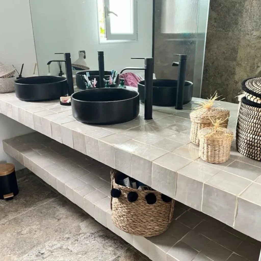 Salle de bain avec zellige gris clair et doubles vasques noires sur plan en pierre naturelle.