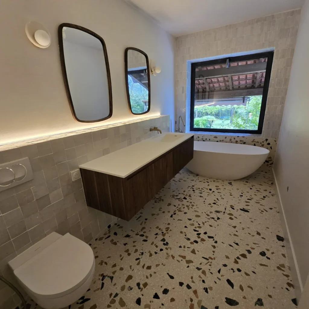 Salle de bain en zellige beige clair avec bois naturel et grande fenêtre, ambiance zen et relaxante.